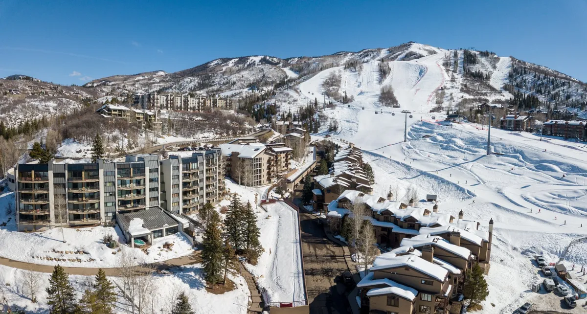 A wide view of snowy mountains, ski slopes, modern buildings, and pine trees. The ski trails appear freshly groomed, and the modern architecture of the buildings reinforces a premium resort ambiance. The setting is lively yet serene, evoking excitement for skiing and the tranquility of a mountain retreat.