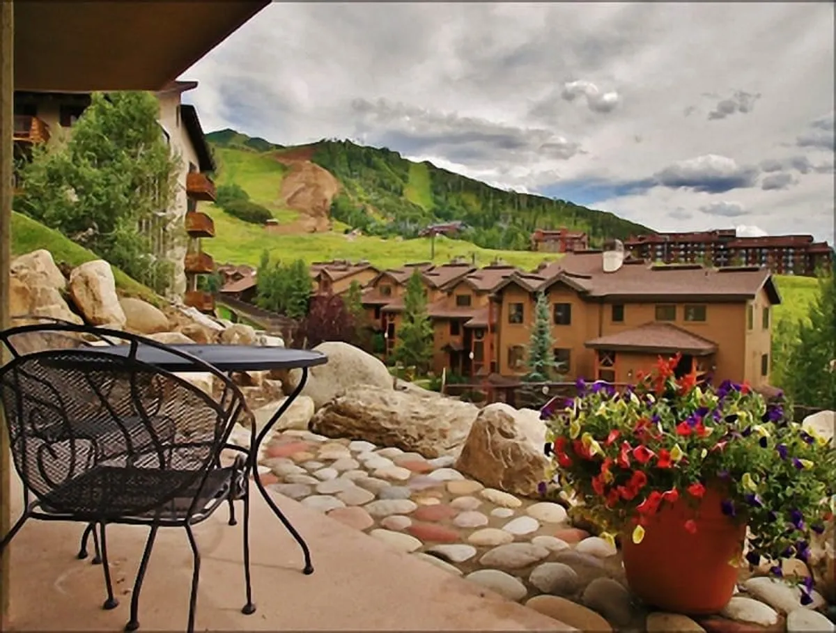 The image shows a cozy outdoor patio with a chair and table set accented by a colorful flower pot, all facing a breathtaking mountain view. The surrounding buildings are visible but do not obstruct the sweeping vista. The scene evokes a tranquil mood, perfect for relaxing outdoors, savoring a morning coffee, or sharing moments with friends as you soak in the natural beauty of Steamboat Springs.