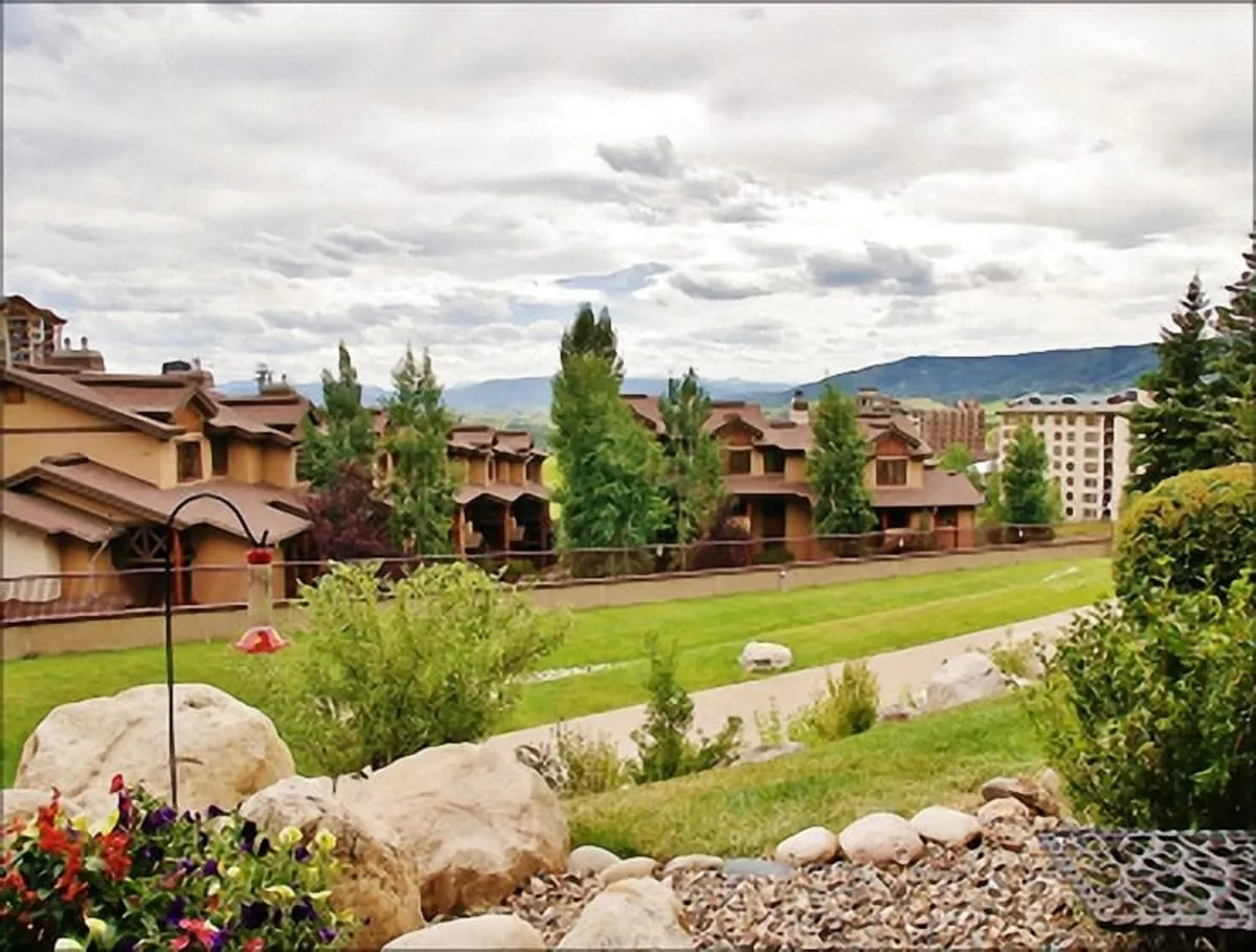 A beautifully landscaped garden with a winding pathway, surrounding buildings in the background, and majestic mountains rising beyond. The scene is bathed in warm sunset light, with vibrant seasonal flower blooms and the possibility of spotting local wildlife. People are depicted walking, enjoying picnics, or simply exploring the natural surroundings.