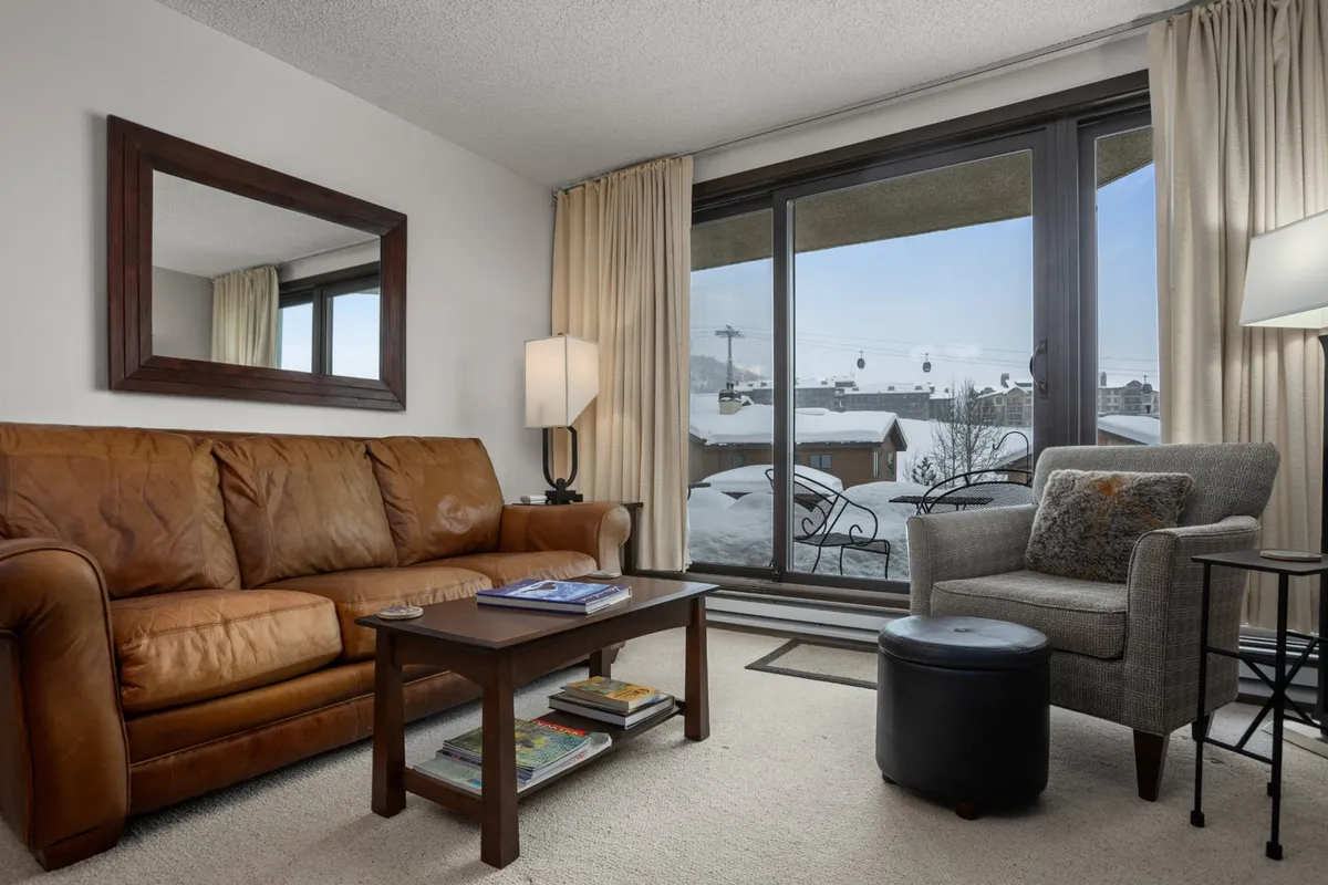 A warm, inviting living room featuring a leather sofa, a coffee table, and an armchair, all arranged for socializing or relaxation. A large window offers a direct view of the surrounding landscape, further emphasizing the connection to nature and mountain scenery.