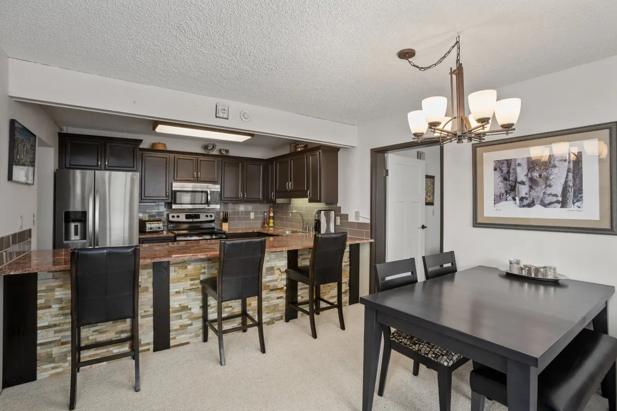 This image centers on the open kitchen and dining area, featuring a striking kitchen island, a large dining table set for groups, and a modern chandelier overhead. The finishes are contemporary with warm touches, and the layout shows clear sightlines between the kitchen and dining area, enhancing the sense of communal gathering.