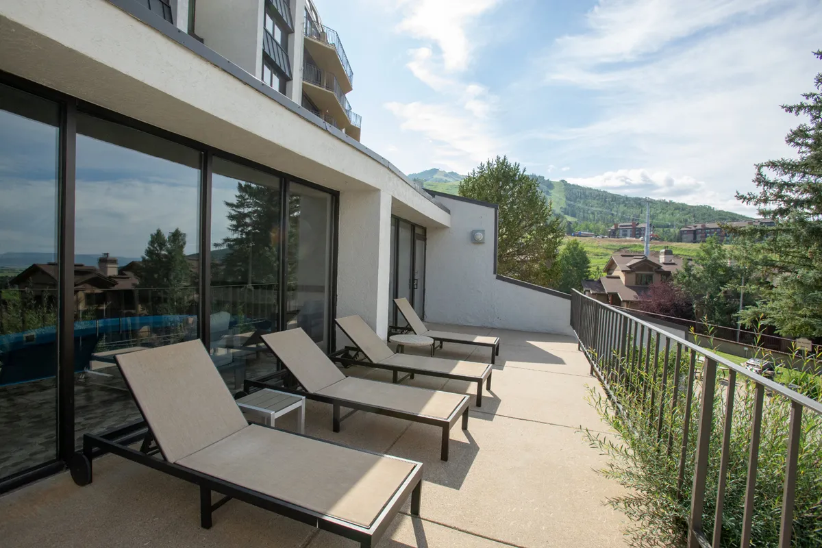 An inviting outdoor lounge area with comfortable chairs positioned to overlook the stunning mountain view. The space is designed for sunbathing, relaxing, and socializing, with large glass windows behind the seating area reflecting the scenic environment.