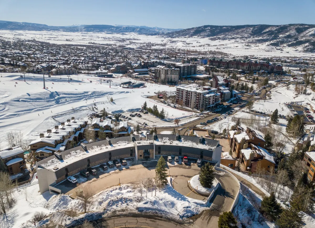 Exterior view of a modern building set against snow-covered mountains and slopes. There are residential buildings, visible ski runs, and architectural details that emphasize both adventure and comfort.