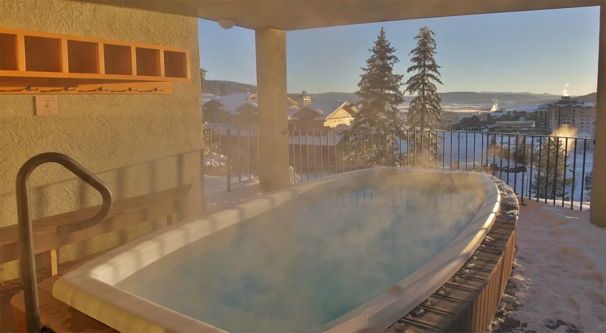 A serene outdoor hot tub steams against a snow-covered landscape, framed by trees and panoramic mountain views. The inviting water contrasts beautifully with the snowy surroundings, suggesting relaxation after a day on the slopes.