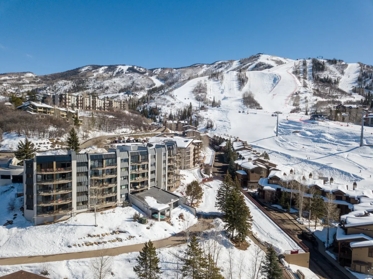 This image captures a modern condominium building nestled beside snow-covered ski slopes, with pine trees and a majestic mountain backdrop. The ski runs are clearly visible, indicating direct access to the slopes from the property, and the building's design aligns with contemporary resort aesthetics.