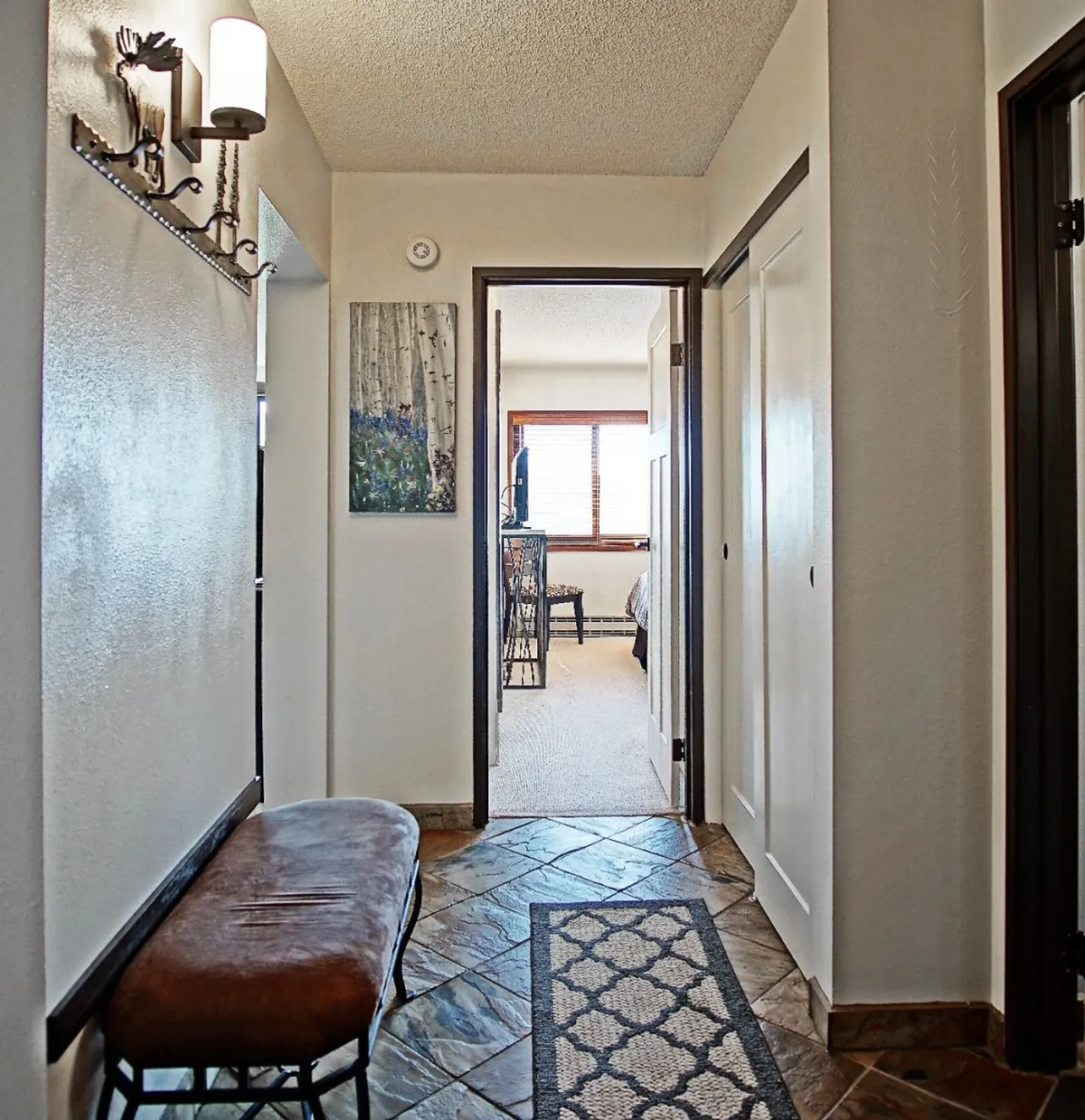An inviting entrance hallway featuring a bench for sitting, tasteful artwork, and a view into an adjacent bedroom. Soft lighting and clean lines set a welcoming tone, providing a smooth entry from the outdoors.