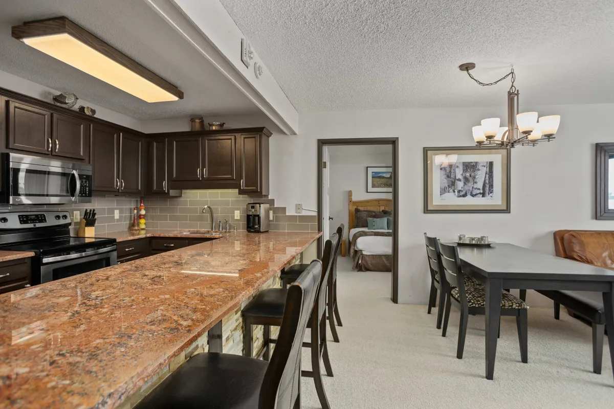 This image provides a different angle that connects the kitchen to the dining area, featuring granite countertops, dark cabinetry, a dining table under a chandelier, and a visible doorway leading to the rest of the home. The warm lighting and setup evoke a welcoming, homey environment ideal for group meals and conversation.