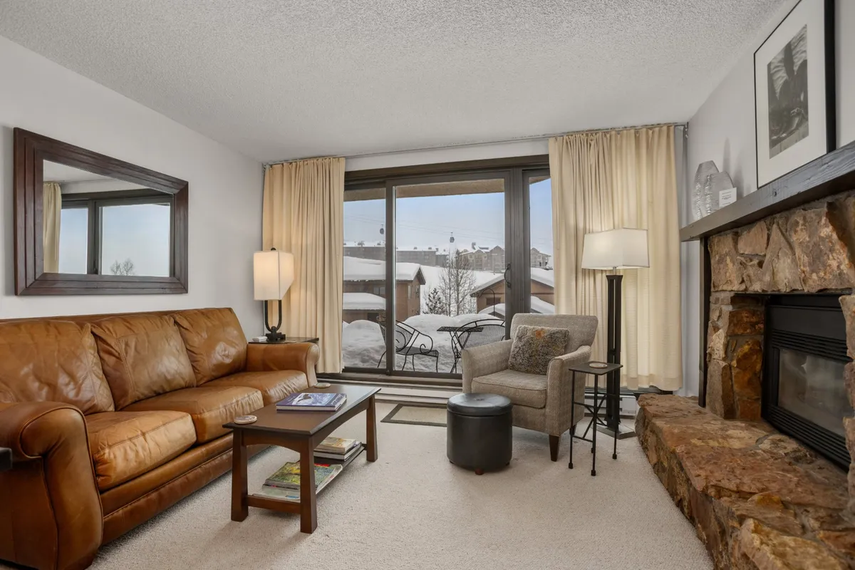 A cozy living room with a plush leather sofa faces a stone fireplace. Large windows frame stunning mountain views, allowing natural light to fill the space and connect guests to the outdoors. The inviting ambiance is perfect for relaxing by the fire, reading, or enjoying the scenery after a day on the slopes.