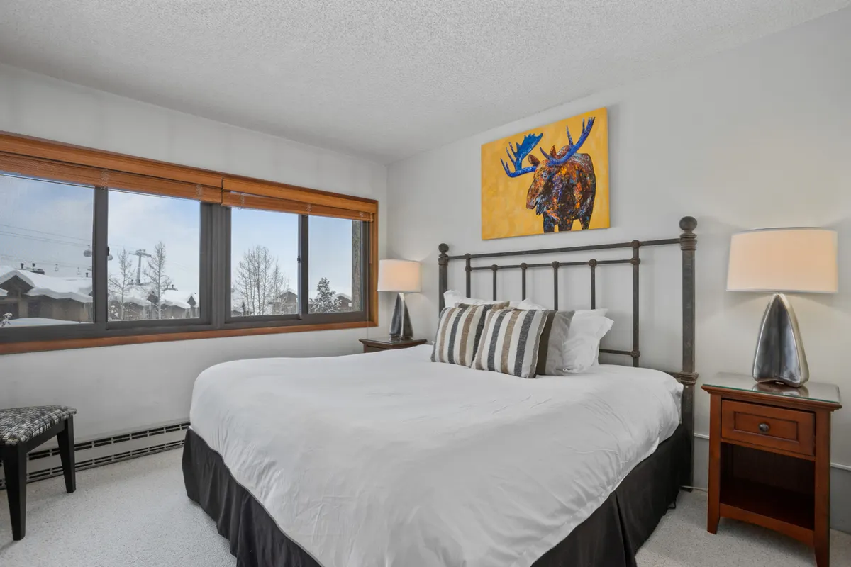 A spacious bedroom featuring a large bed with crisp white bedding, soft pillows, and moose artwork above the headboard. The room is illuminated by natural light from a window framing a peaceful snowy landscape. Nightstands with lamps provide ambient lighting, creating a tranquil setting ideal for relaxation and rest.